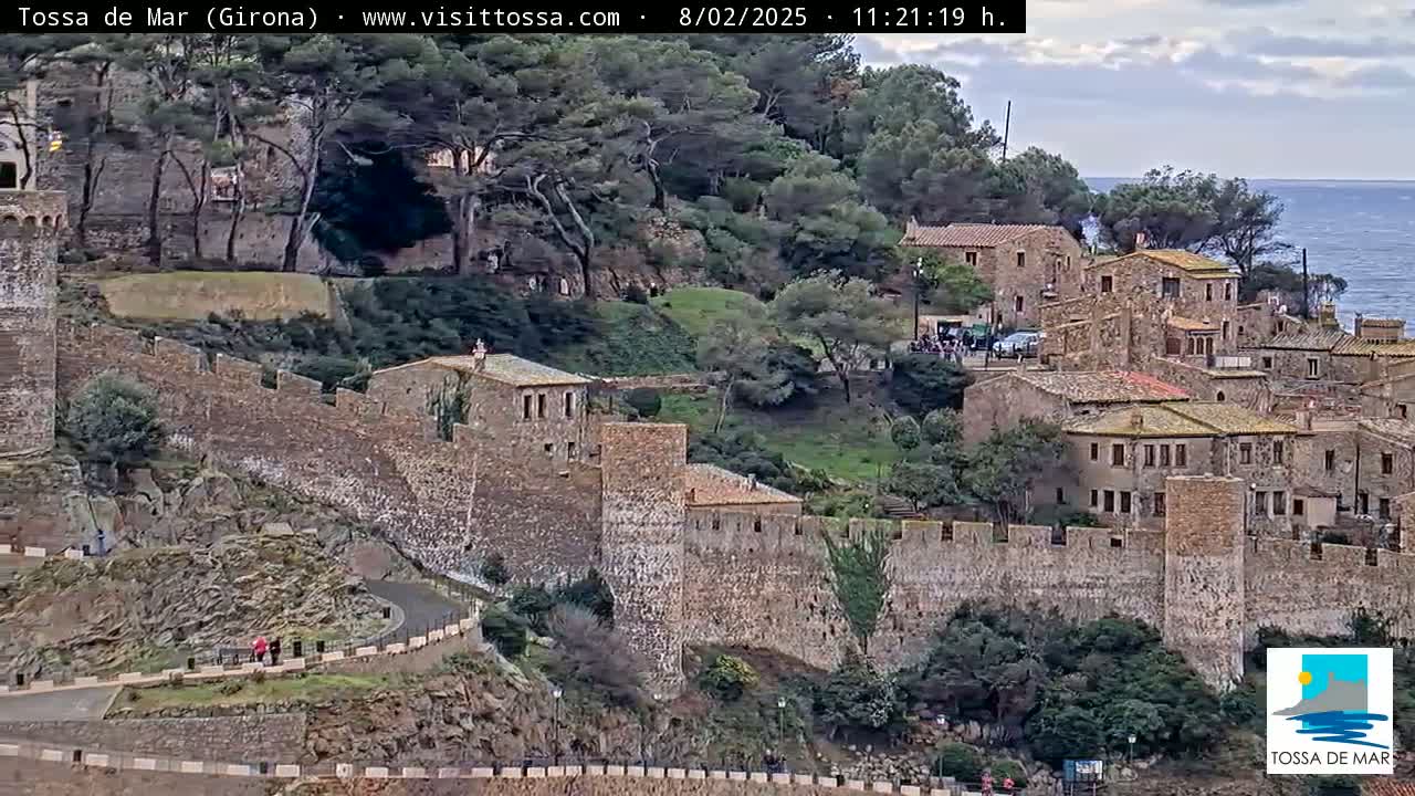 A stone walled medieval town overlooks a sea under a partly cloudy sky.