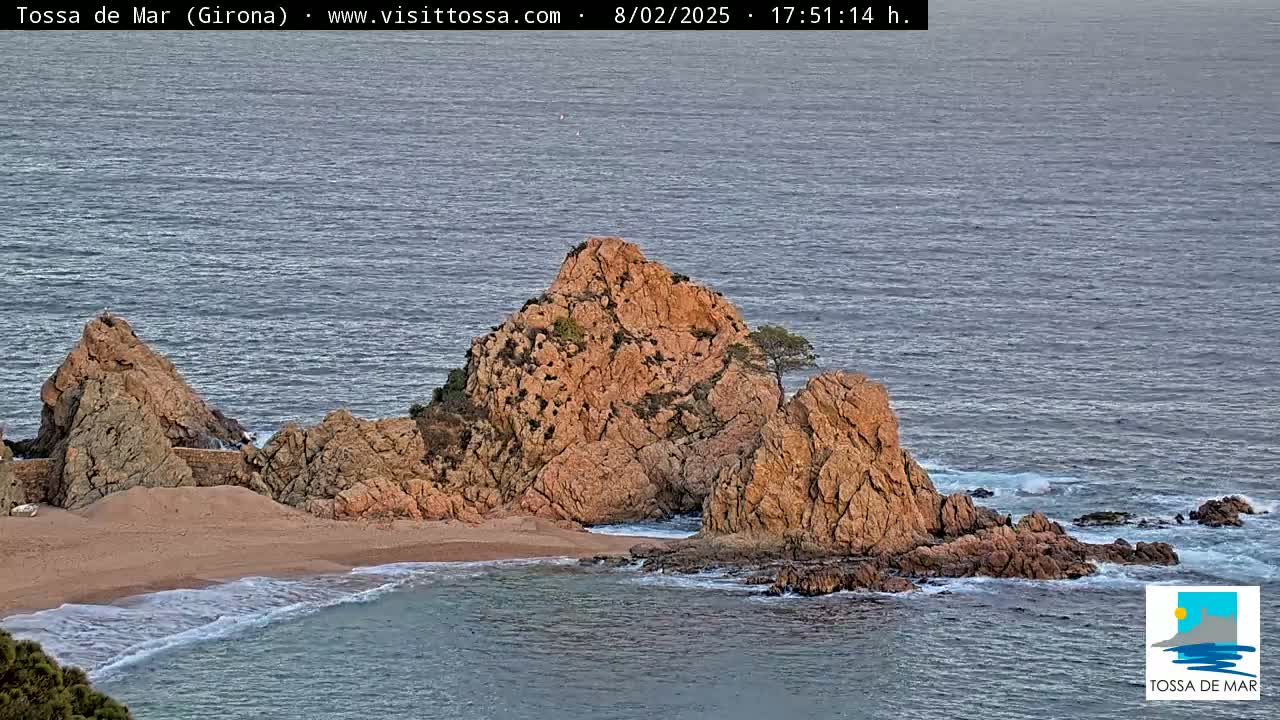 A small, sandy cove nestled between reddish-brown rocky outcrops on a calm sea under a mostly clear sky.