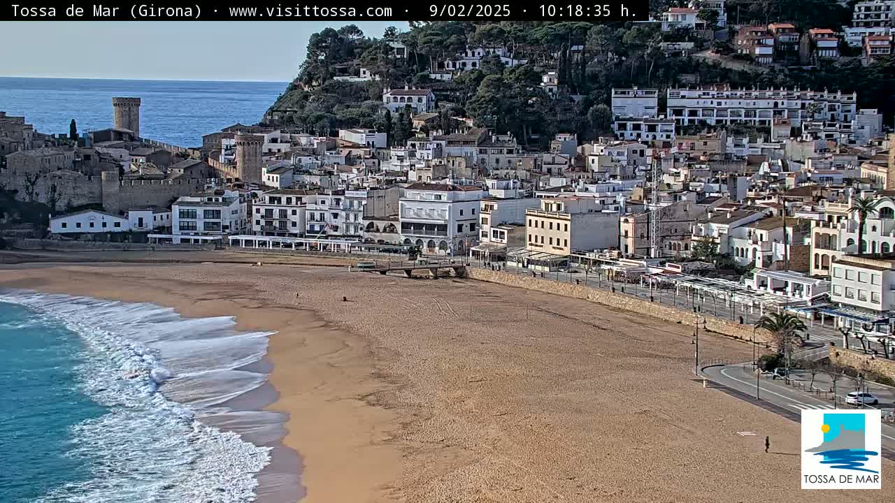 A sandy beach with gentle waves laps the shore in front of a coastal town featuring a medieval castle and numerous buildings that climb a hillside.