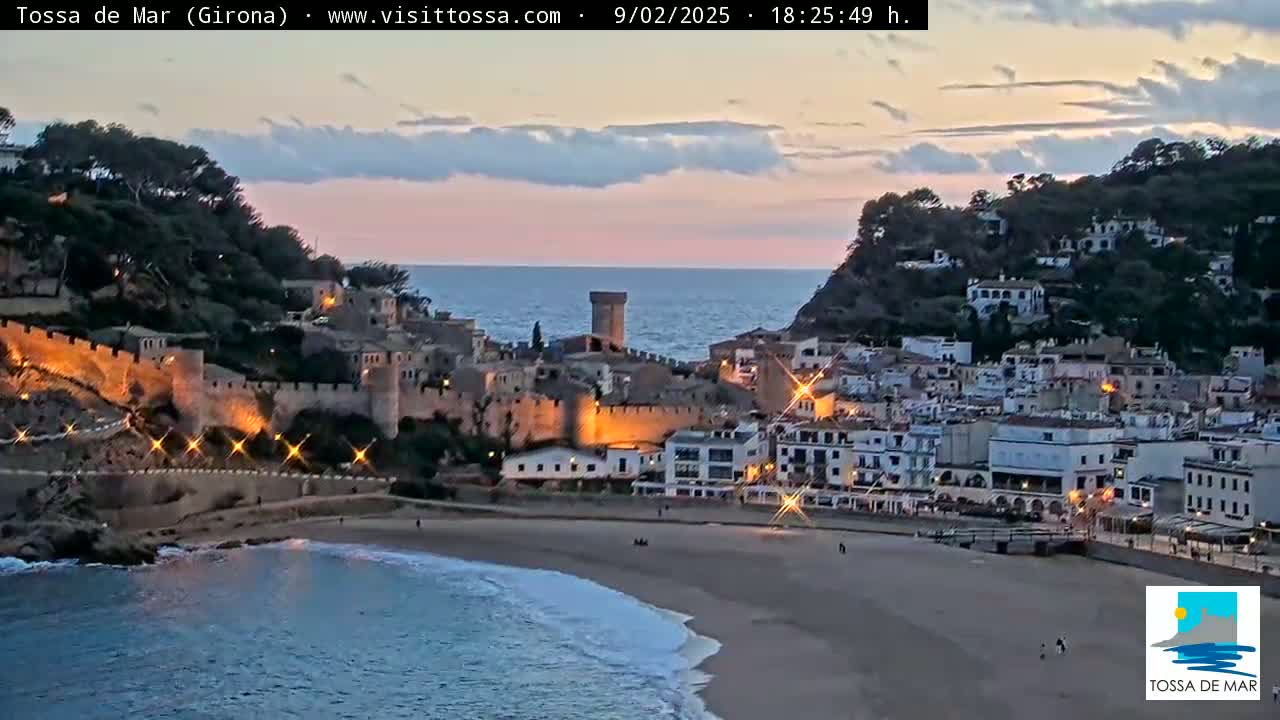 A coastal town with a medieval fortress sits on a hill overlooking a sandy beach and calm ocean at sunset.