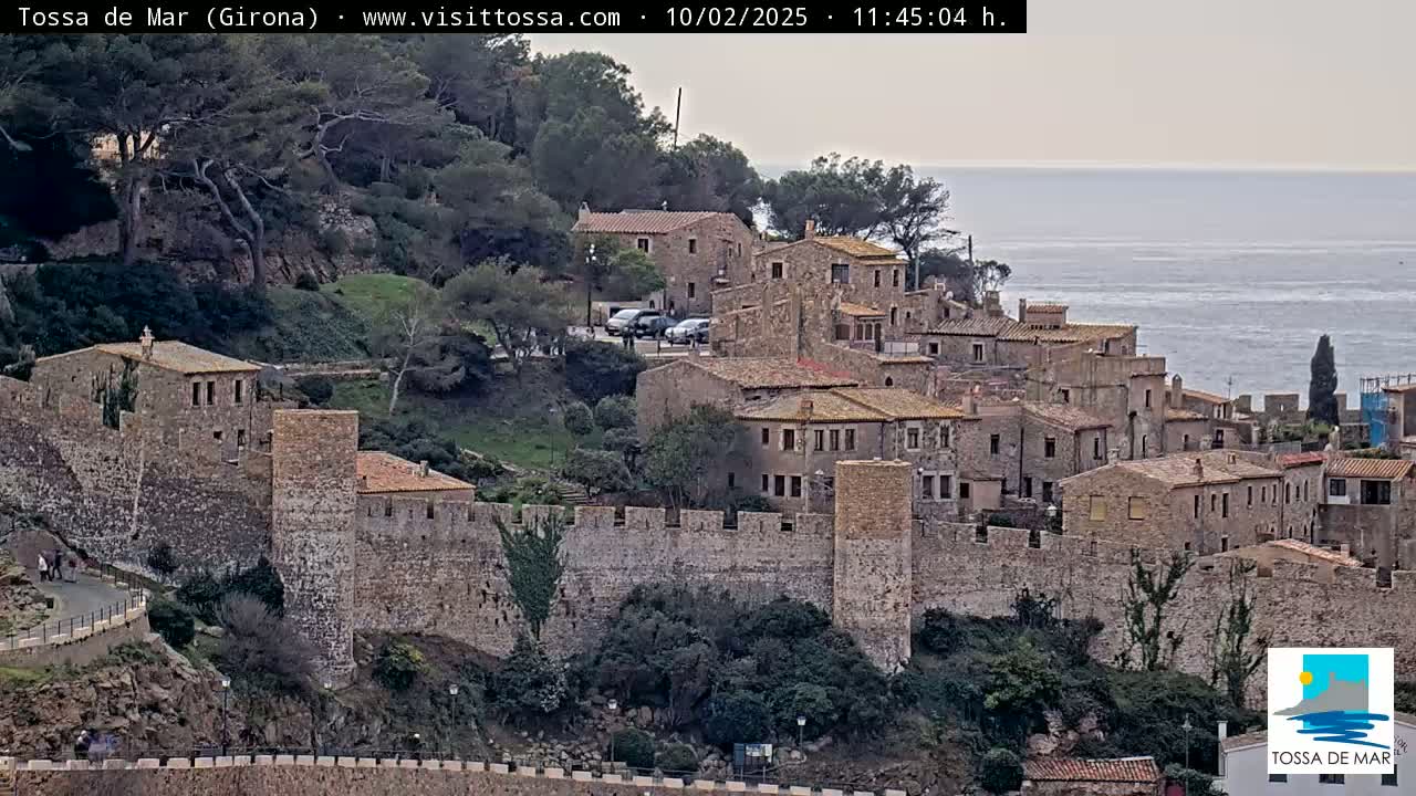 A coastal village with stone buildings and walls overlooks a calm sea under an overcast sky.