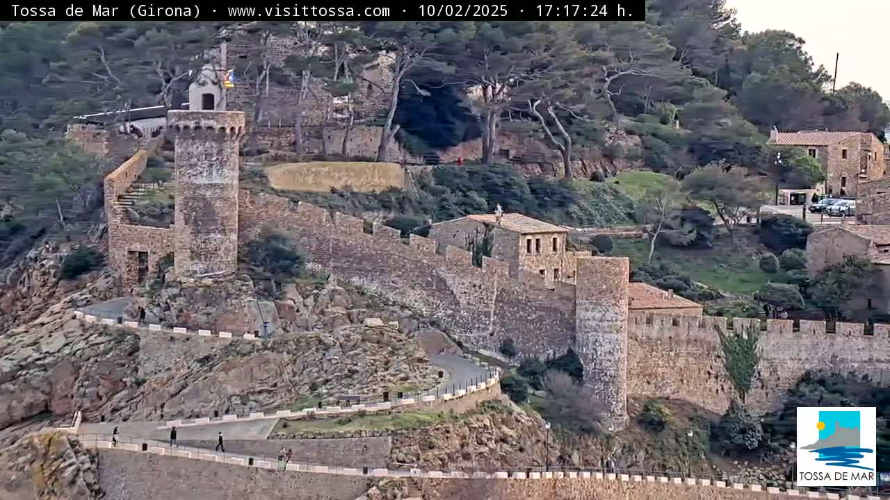 A stone wall and tower overlook a town nestled among green hills and trees on a partly cloudy day.