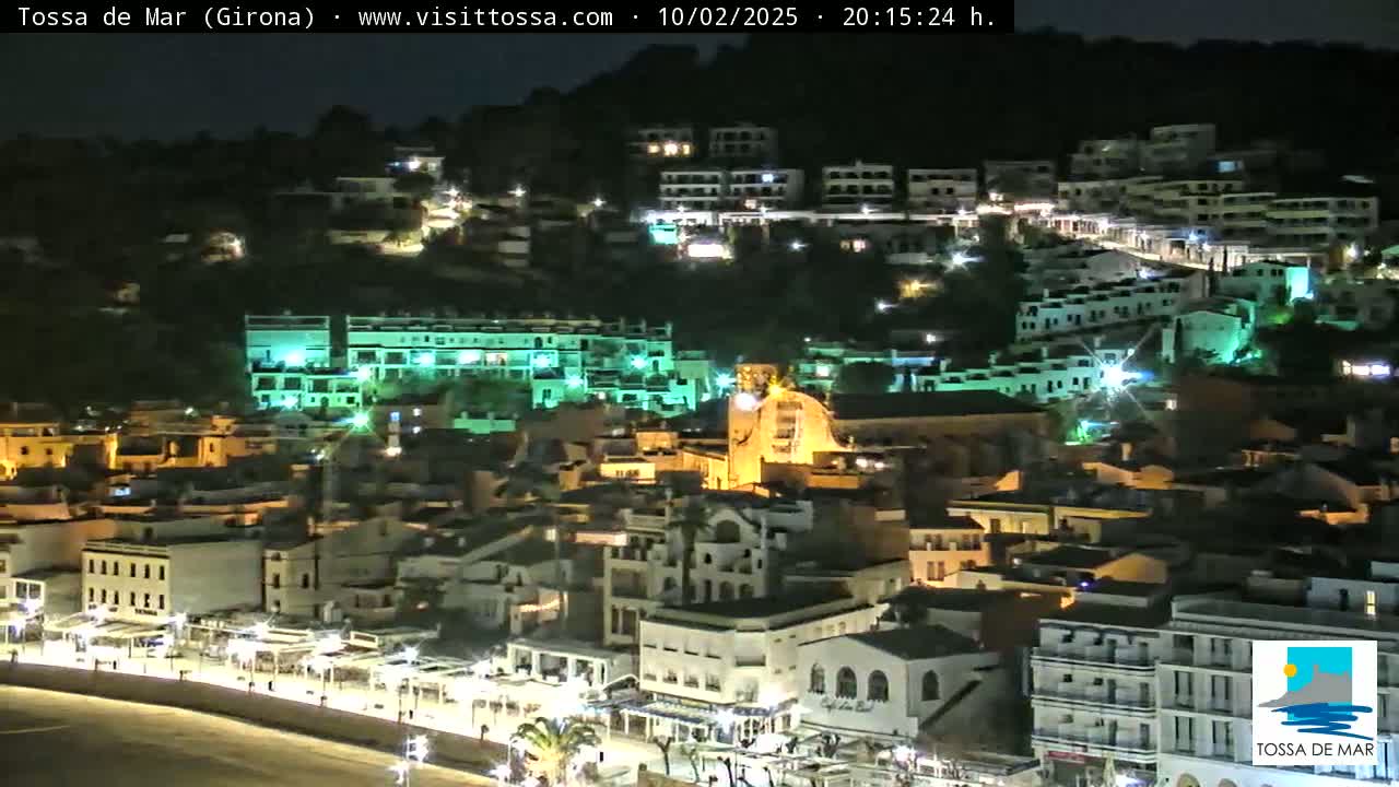 A nighttime view of a coastal town shows buildings illuminated along a hillside, extending down to a beach.