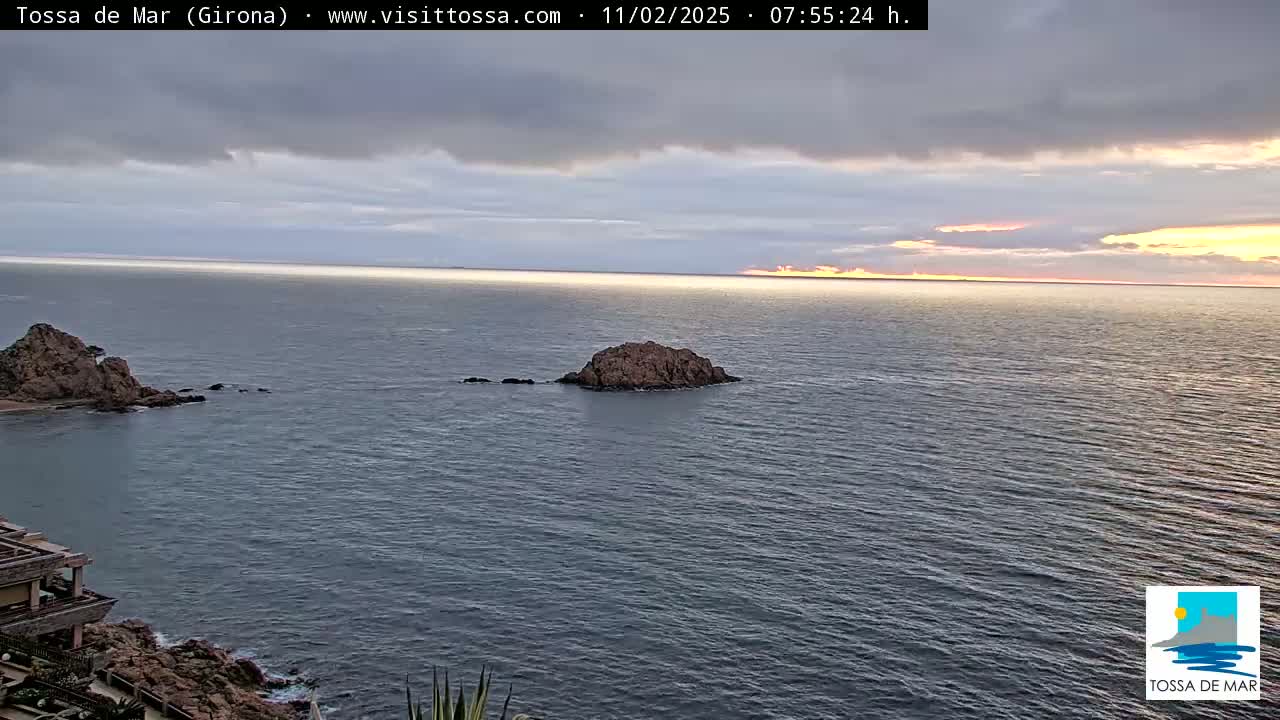A calm sea at sunrise is seen from a coastal viewpoint, with rocky outcrops and a building visible in the foreground.