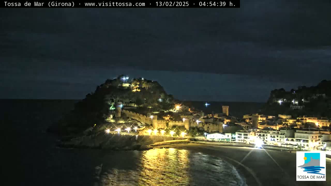 A coastal town at night, illuminated with lights, is situated on a hill overlooking a dark ocean under a mostly clear night sky.