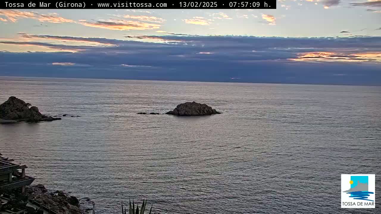 A calm sea under a mostly cloudy sky with hints of sunrise or sunset colors shows two rocky outcrops in the water.