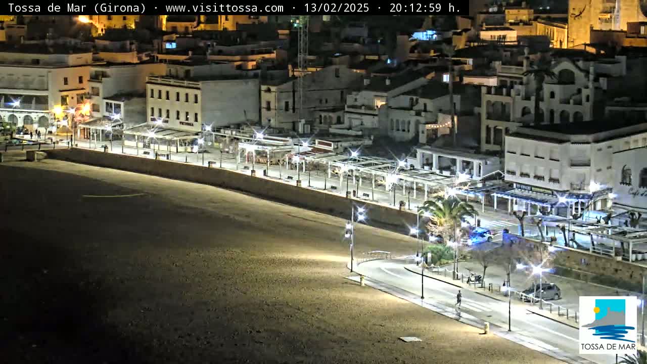 A nighttime, brightly-lit beachside promenade and town with a sandy beach, lined with buildings, palm trees, and streetlights.