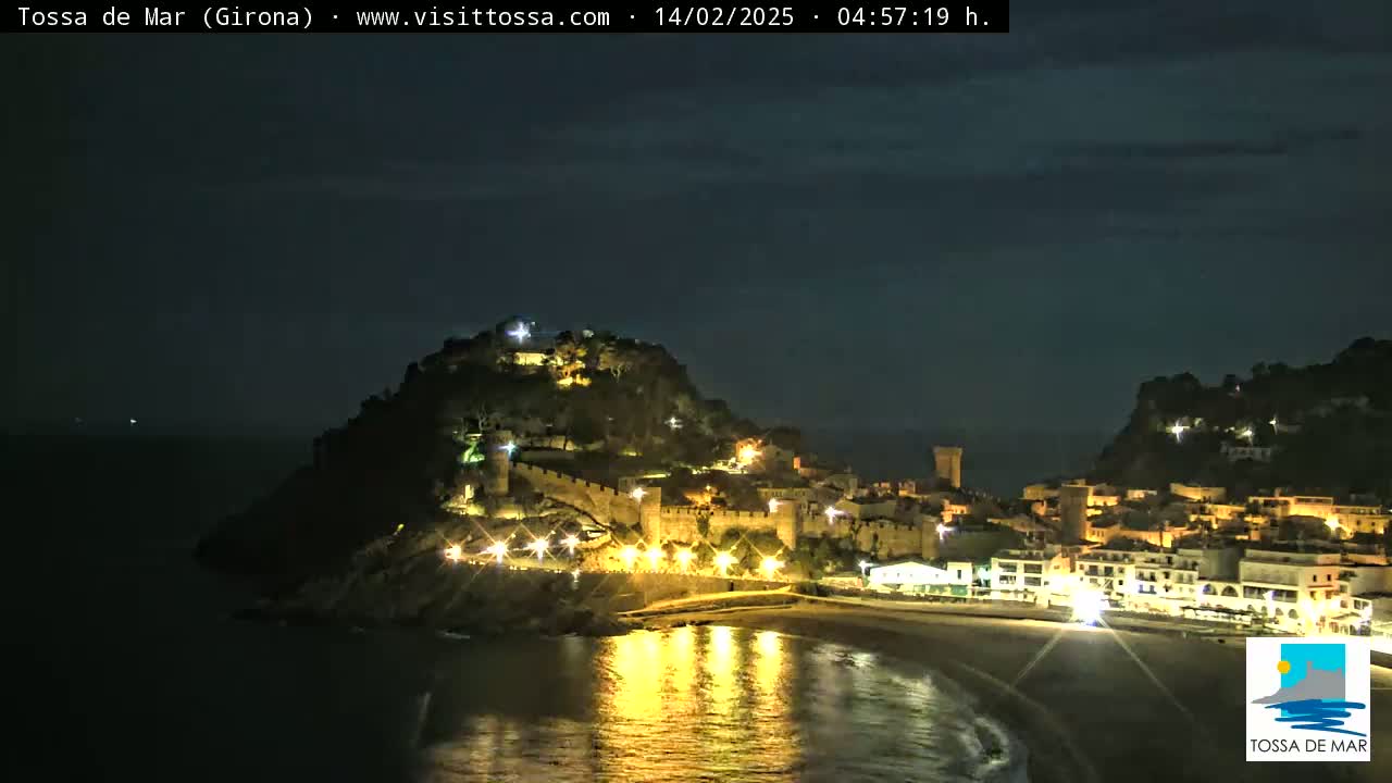 A nighttime coastal town, illuminated, is nestled against a dark hill overlooking a calm, dark sea reflecting the town's lights.