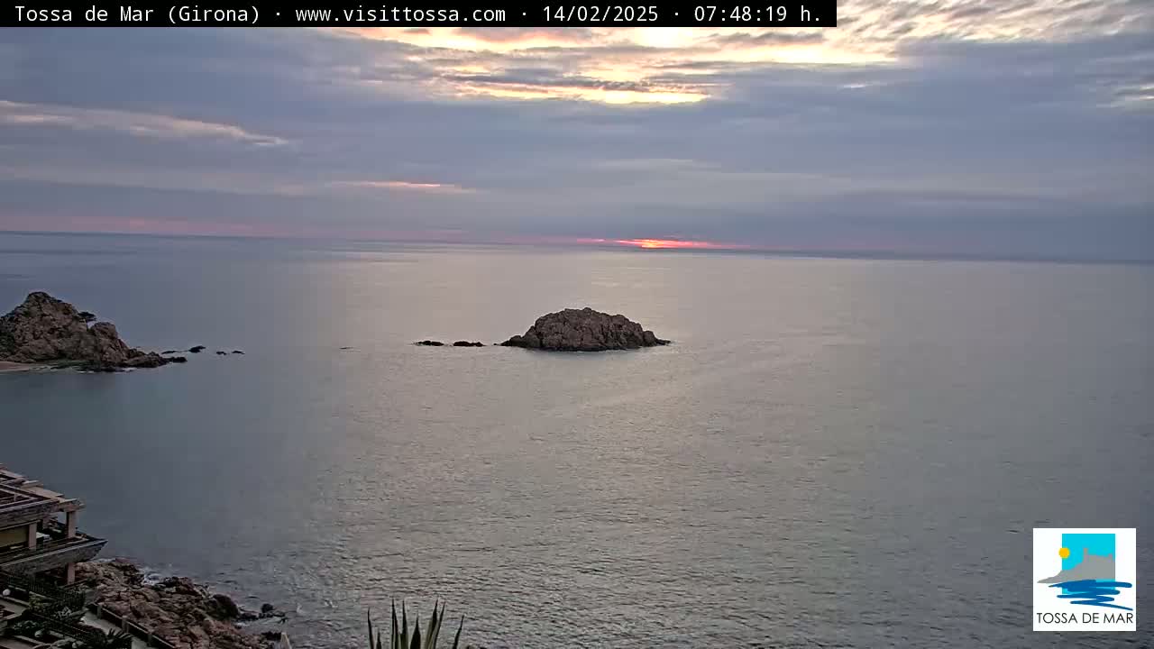 A rocky coastline and small island are visible in a calm sea during a sunrise with cloudy skies.