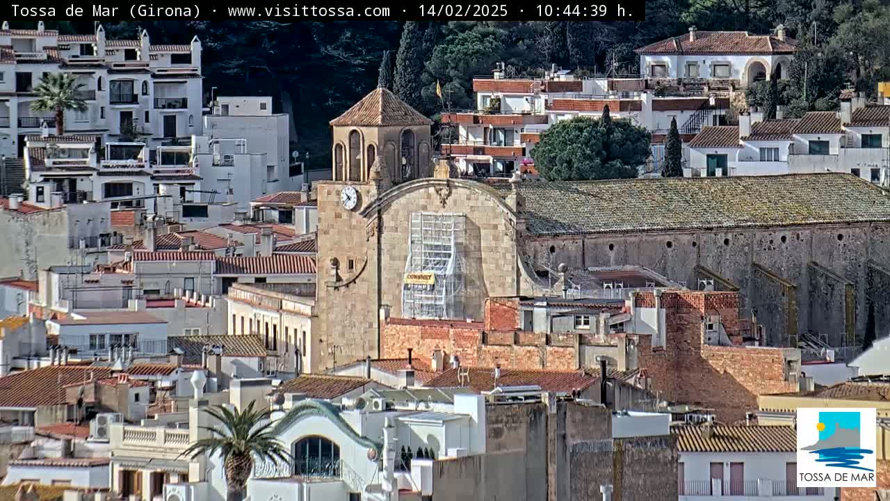 A sunlit coastal town with a stone church undergoing renovations is nestled amongst many white and terracotta-roofed buildings.