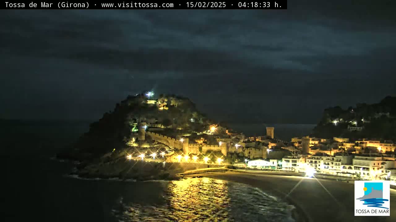 A nighttime coastal town, illuminated by lights, is situated on a hill overlooking a dark ocean under a cloudy sky.