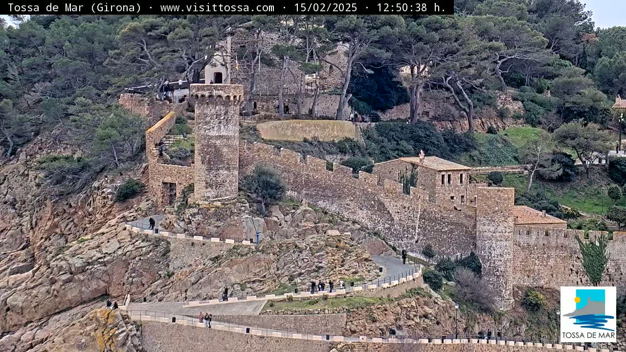 A stone castle wall winds along a rocky hillside dotted with trees and a few people walking on paths, under a clear sky.