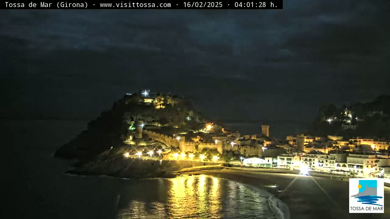 A nighttime view of a coastal town, illuminated with lights, nestled against a hill under a dark, cloudy sky.