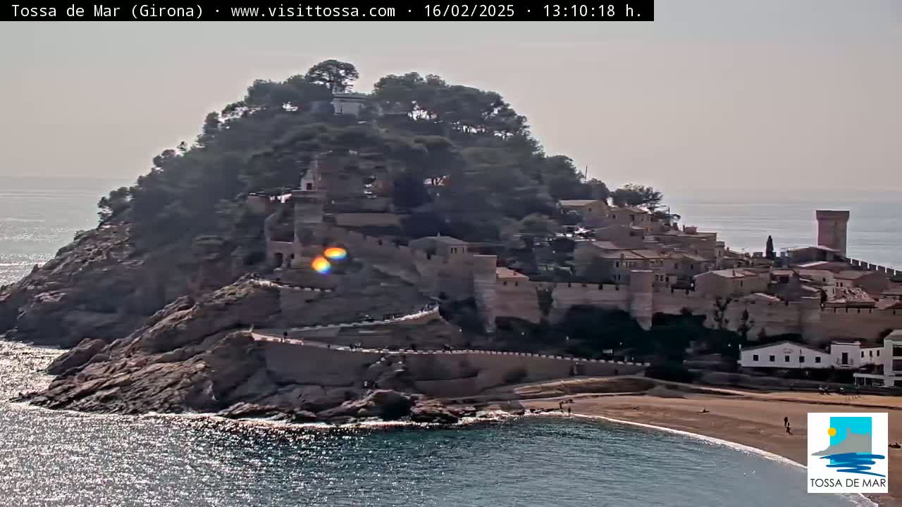 A coastal town with medieval-style walls and buildings sits atop a rocky hill overlooking a calm, sunlit sea.