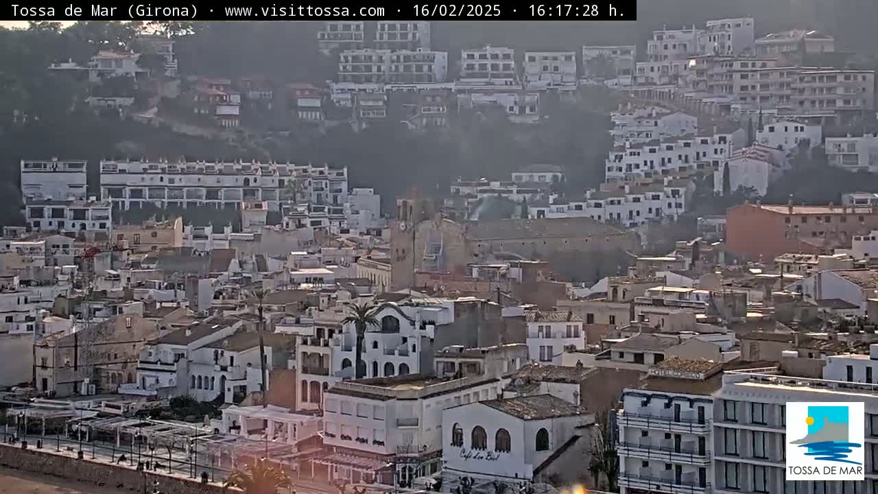 A hazy, sunny day overlooks a dense collection of buildings in a coastal town, nestled against a hillside.