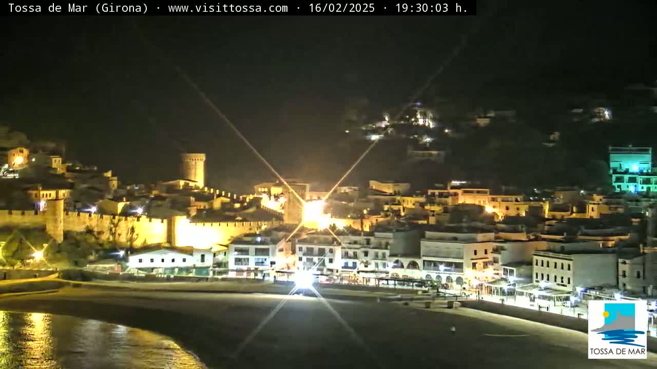 A nighttime view of a coastal town, illuminated, with a beach and calm water in the foreground.
