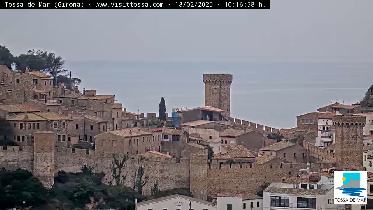 A stone walled medieval town sits on a cliff overlooking a calm sea under an overcast sky.