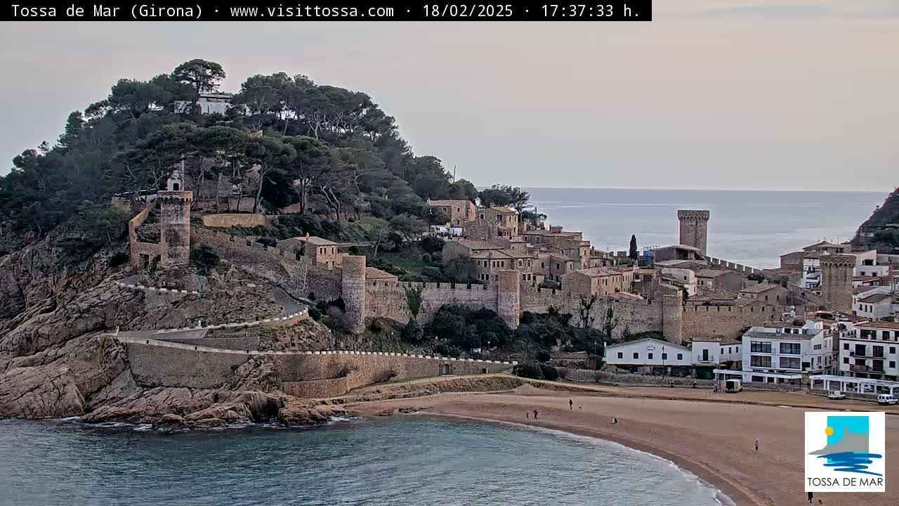 A medieval walled town sits on a cliff overlooking a calm, sandy beach and a tranquil sea under an overcast sky.