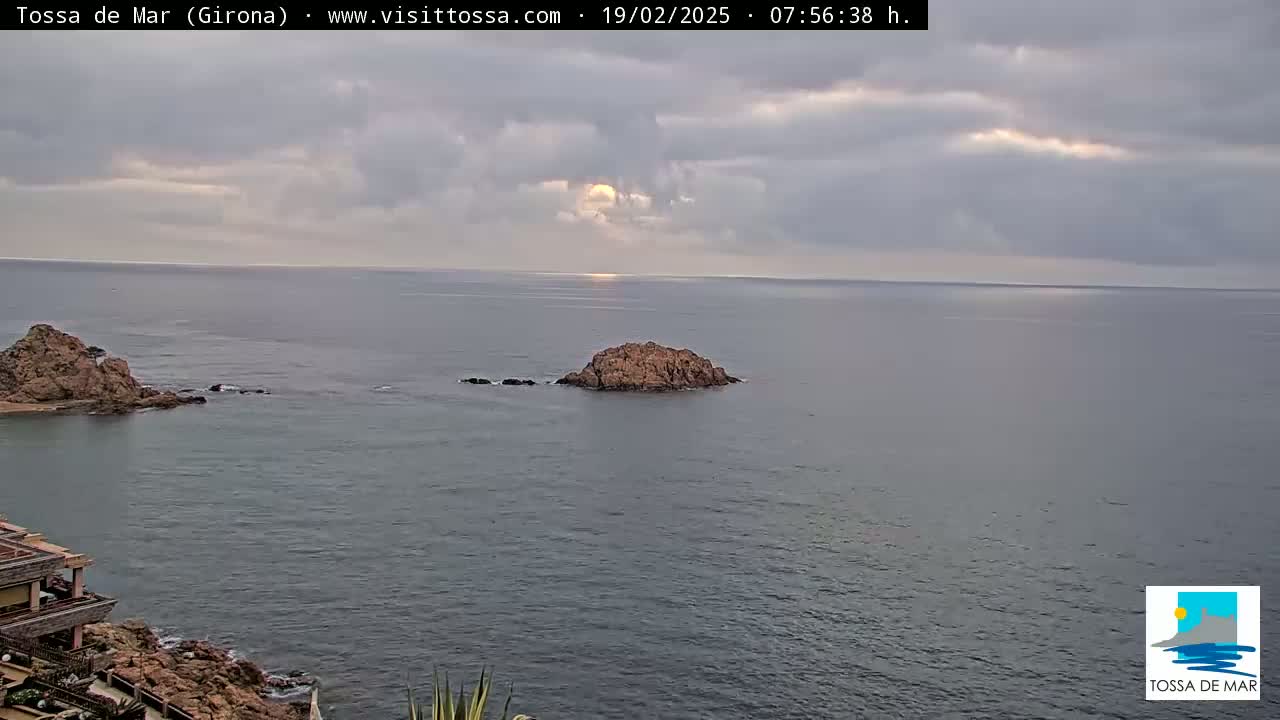 A rocky coastline with a small island in a calm sea under a partly cloudy sky.