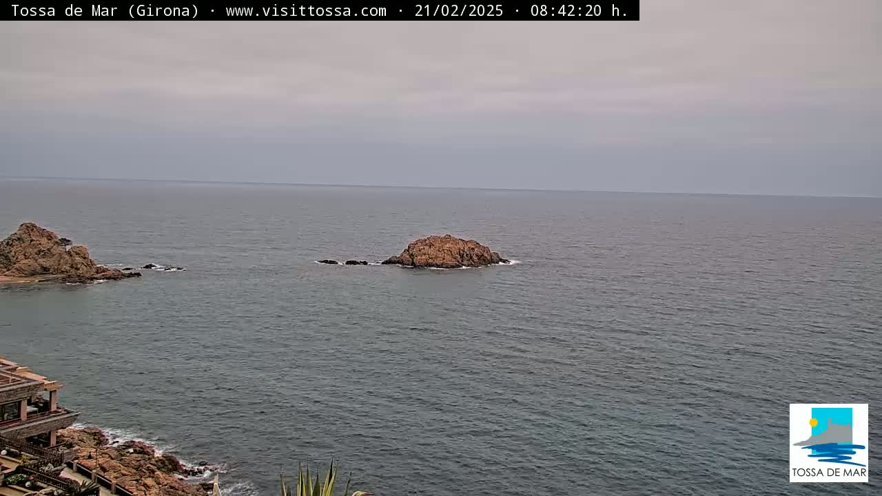 A rocky outcrop sits in a calm, gray sea near a rocky coastline, under an overcast sky.