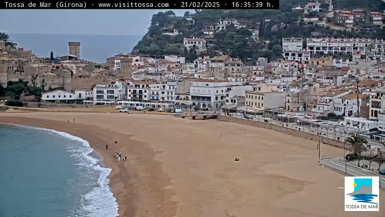 A sandy beach with a few people on it borders a town of white buildings nestled against a hillside under an overcast sky.