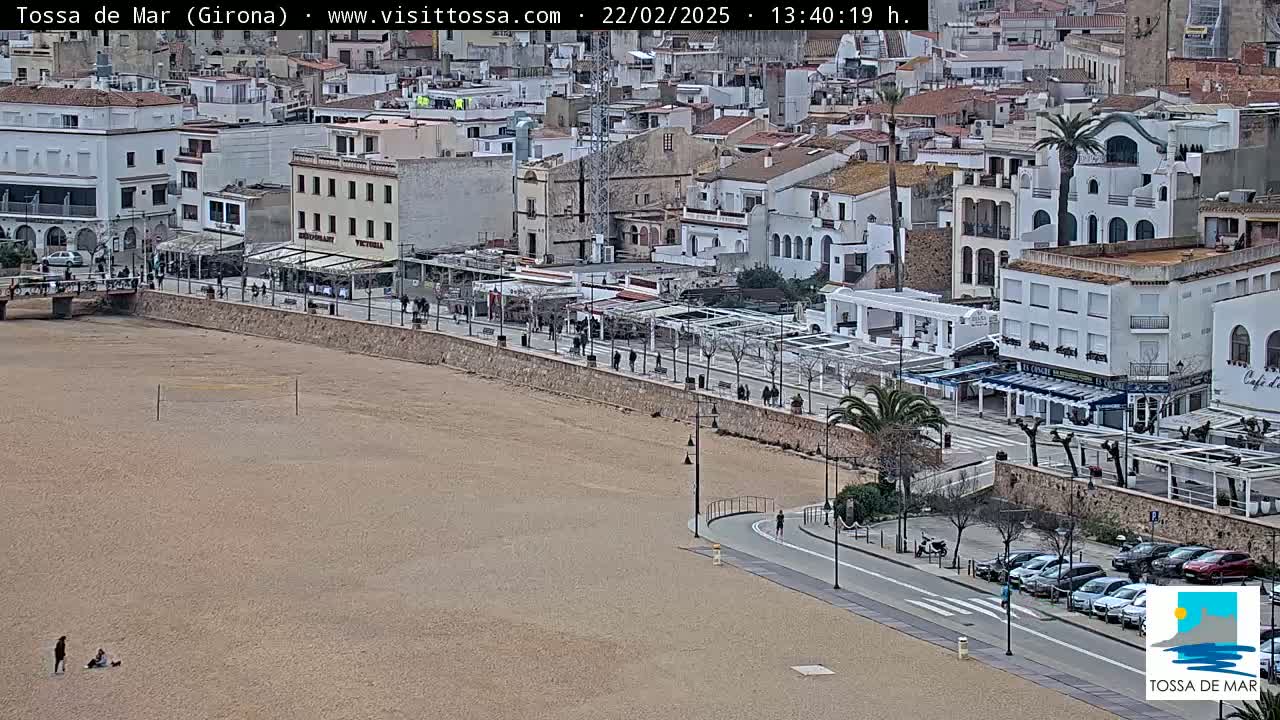 A mostly empty sandy beach stretches below a seaside town on a partly sunny day.