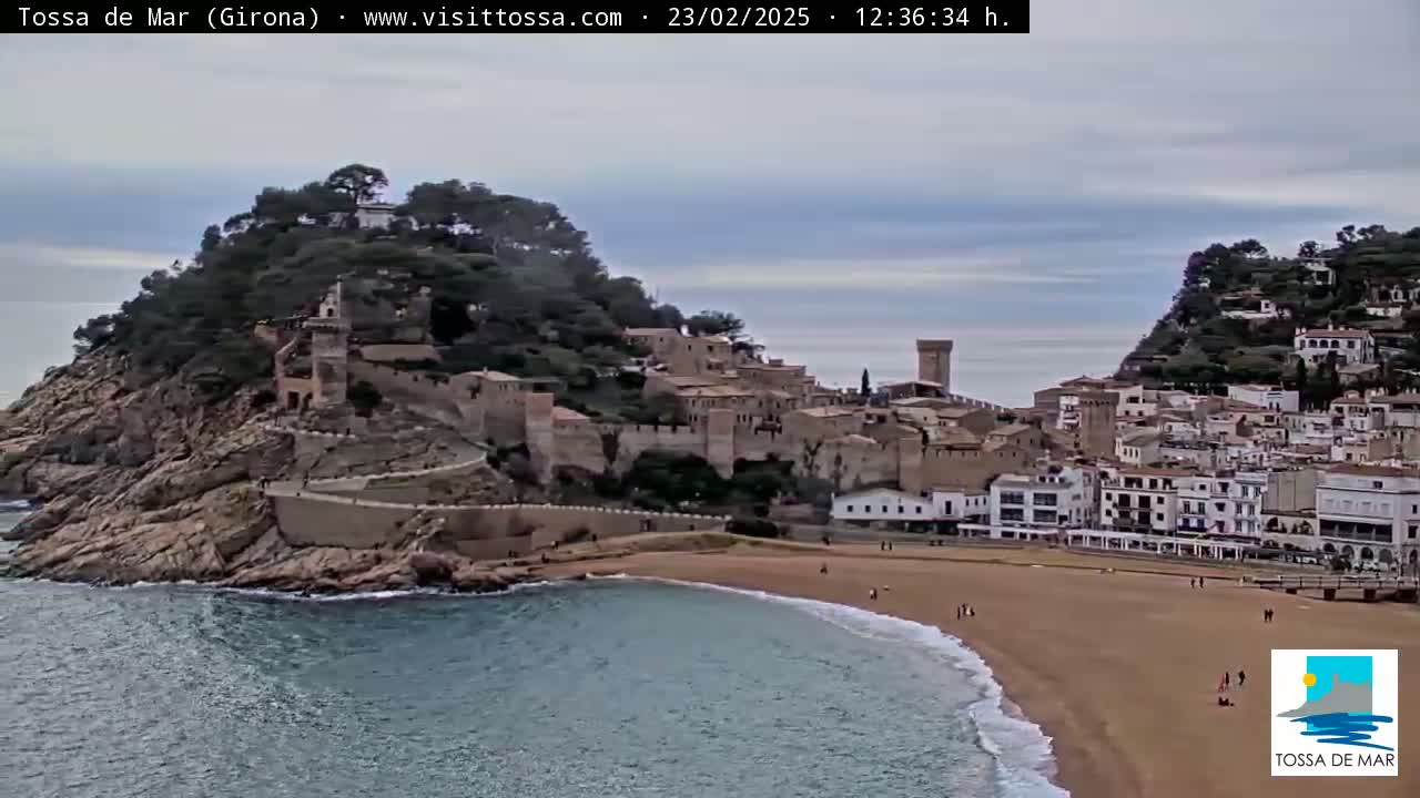 A coastal town with a medieval castle perched atop a cliff overlooking a sandy beach and calm ocean under an overcast sky.