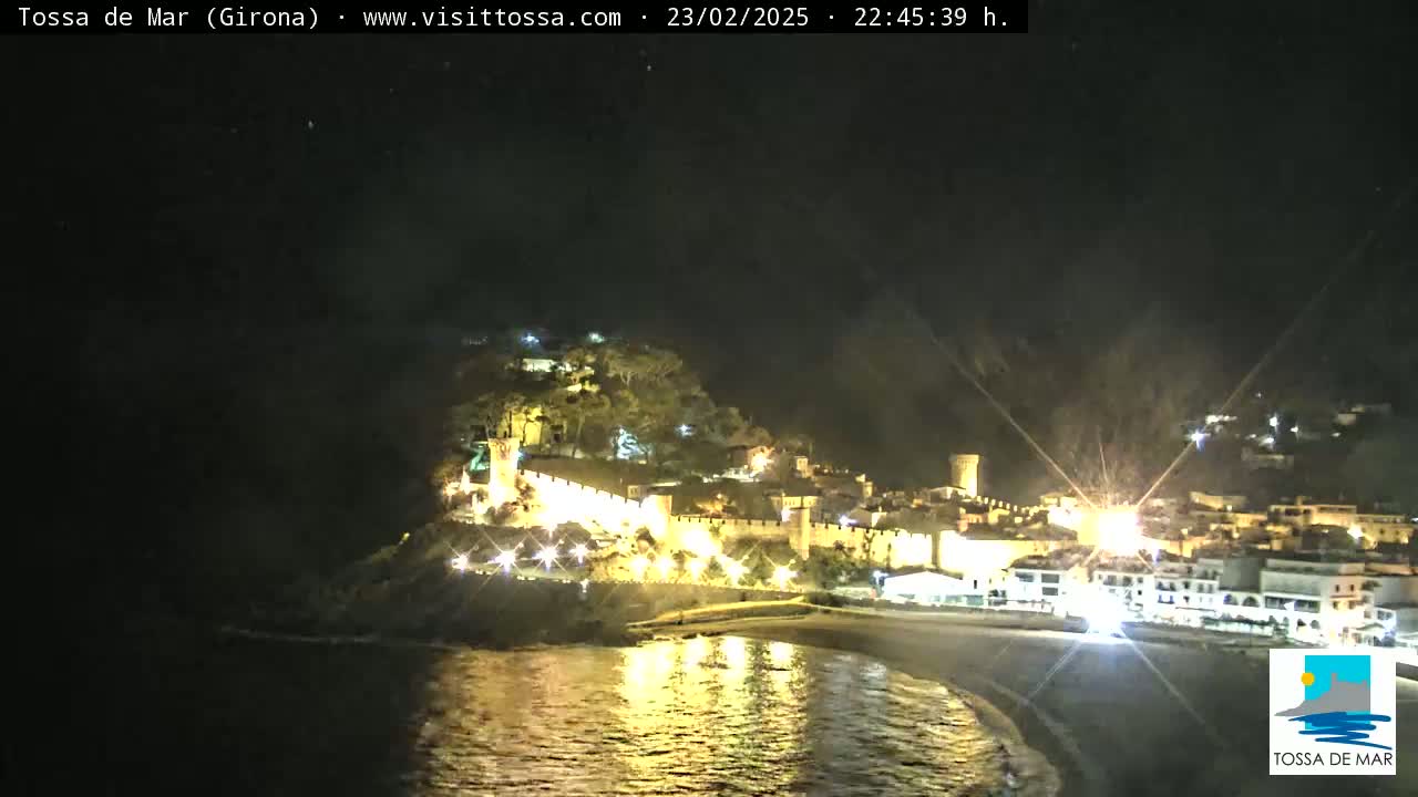 A nighttime view of a coastal town with a walled fortress illuminated, overlooking a calm bay.