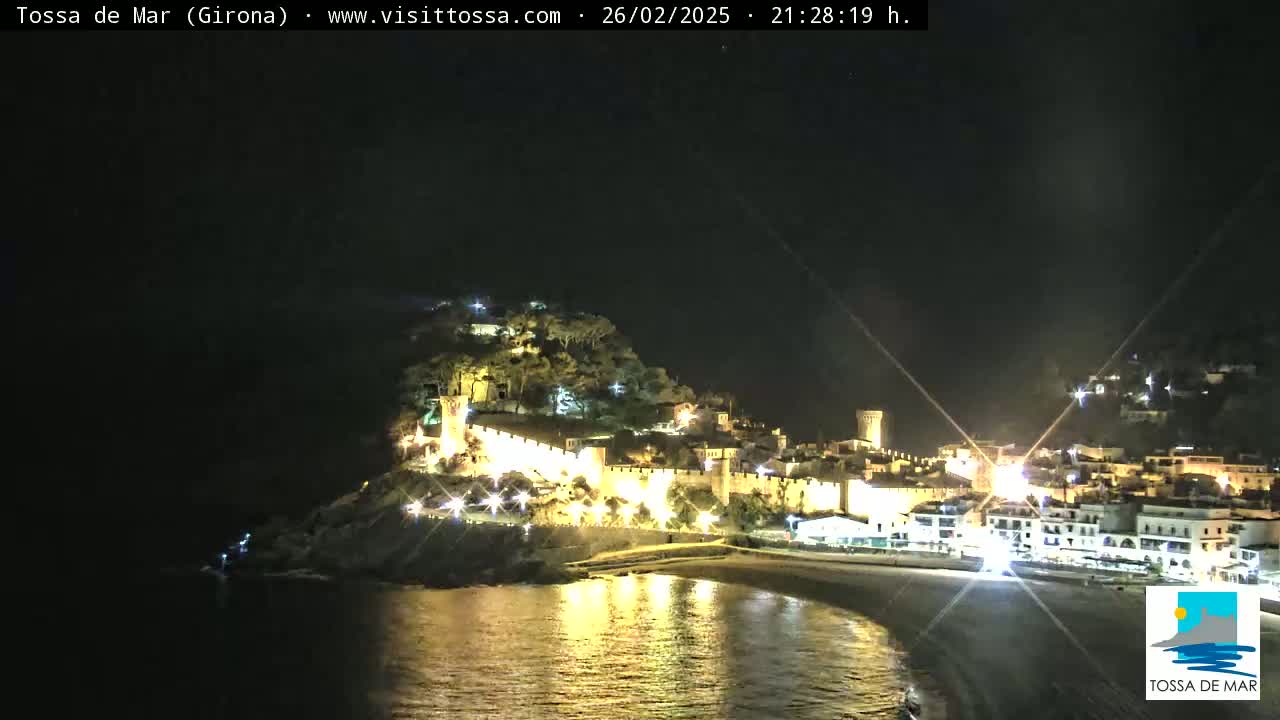 A nighttime coastal town with a walled fortress and a beach, illuminated by lights, under a clear, dark sky.
