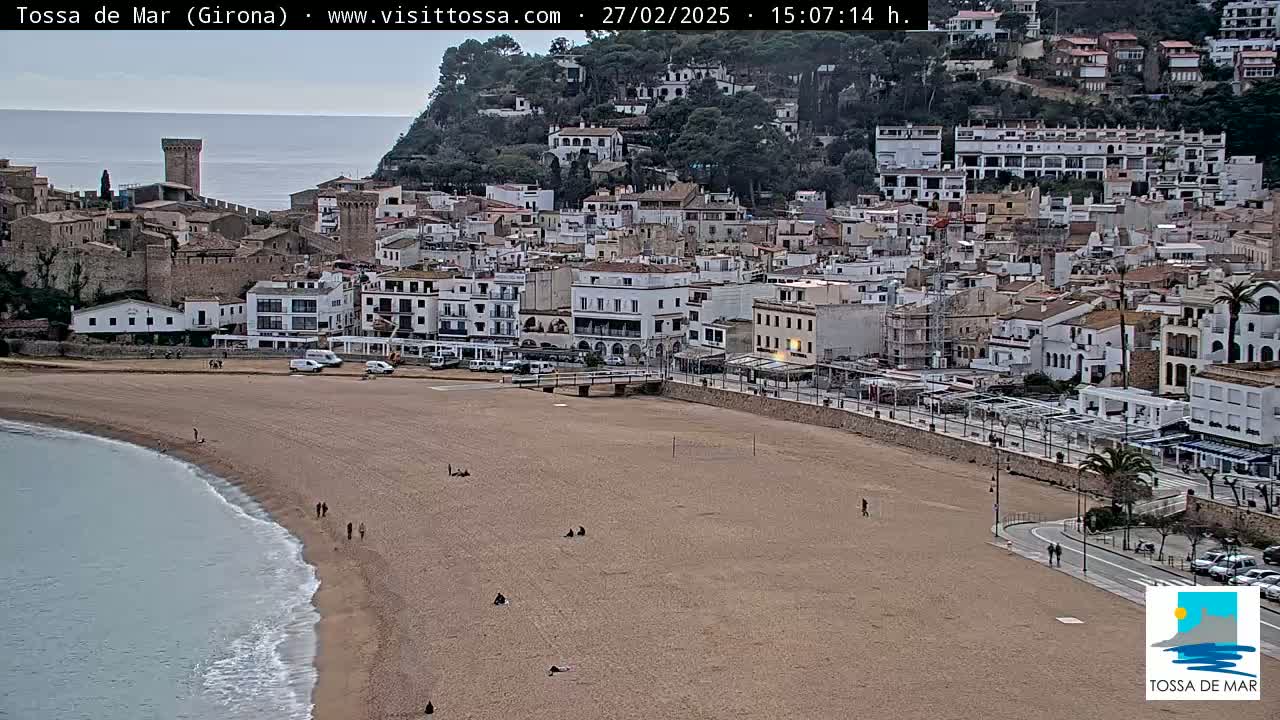 A sandy beach with a few people on it curves into a calm sea beside a town of white buildings nestled against a hillside under an overcast sky.