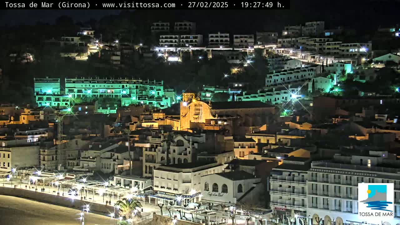 A nighttime view of a coastal town, with buildings lit up along a waterfront promenade.