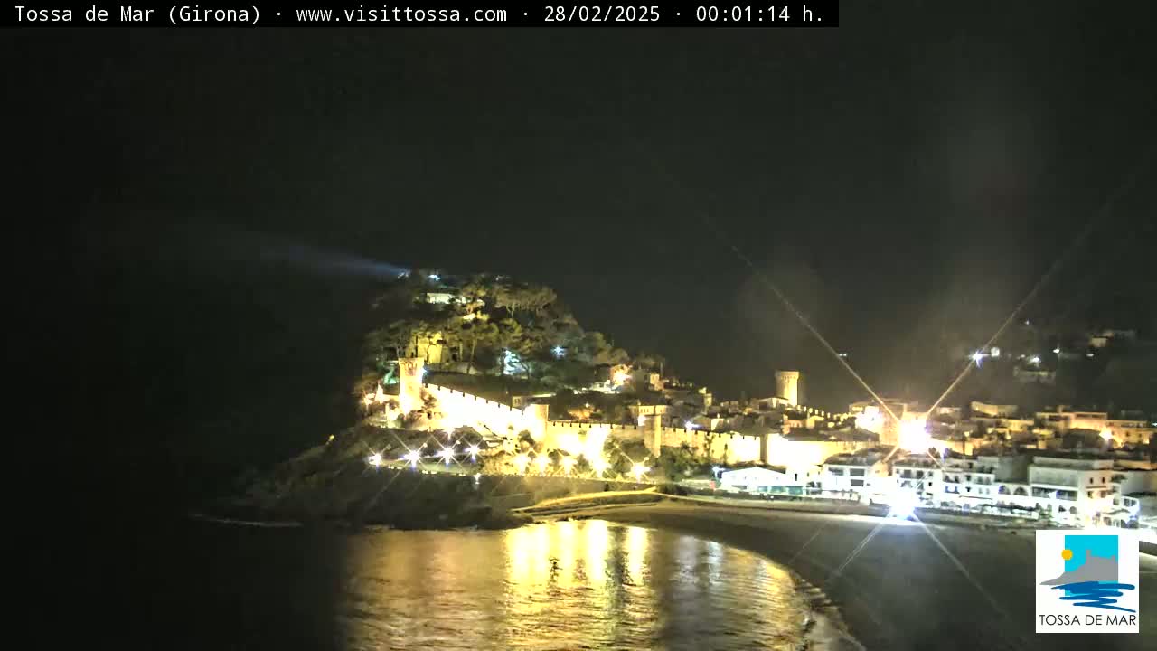 A nighttime view of a coastal town with a walled fortress, illuminated buildings, and a calm body of water reflecting the lights.