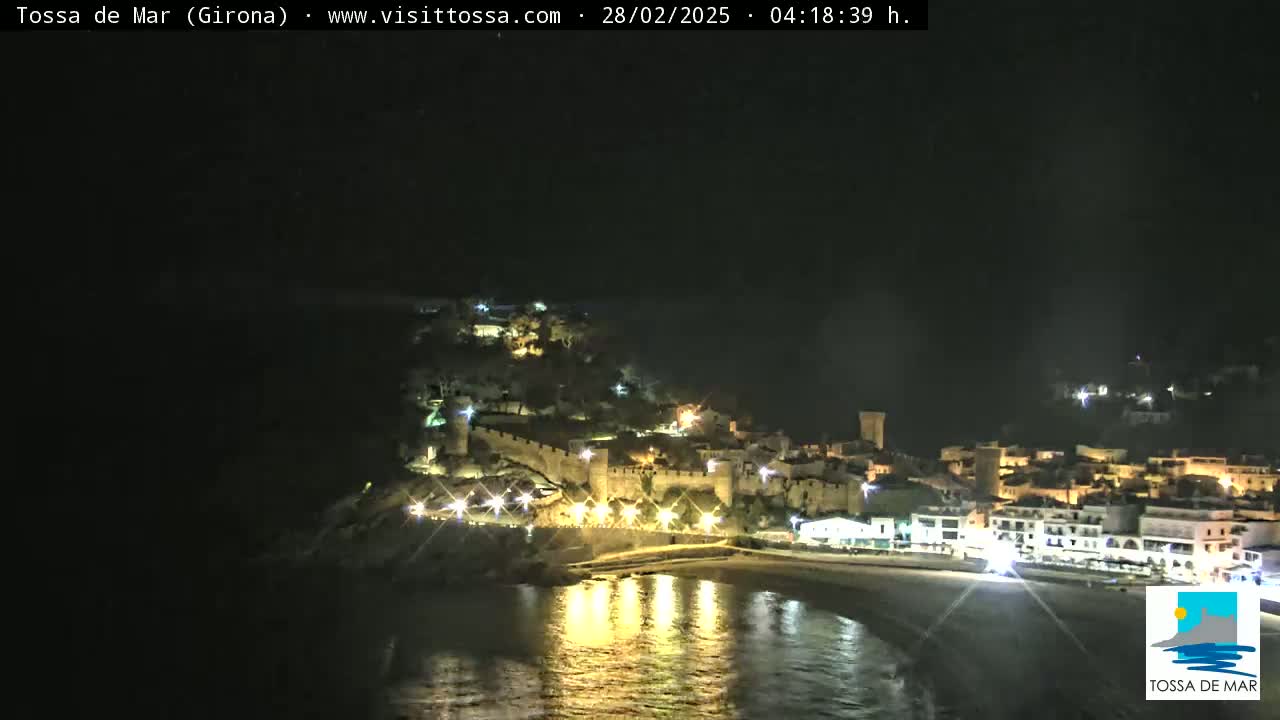 A nighttime view of a coastal town with a castle, illuminated by lights, reflecting on calm water.