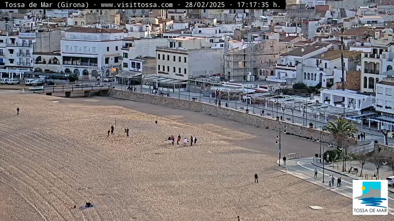A sandy beach with a few people on it lies below a town of white buildings under a clear sky.