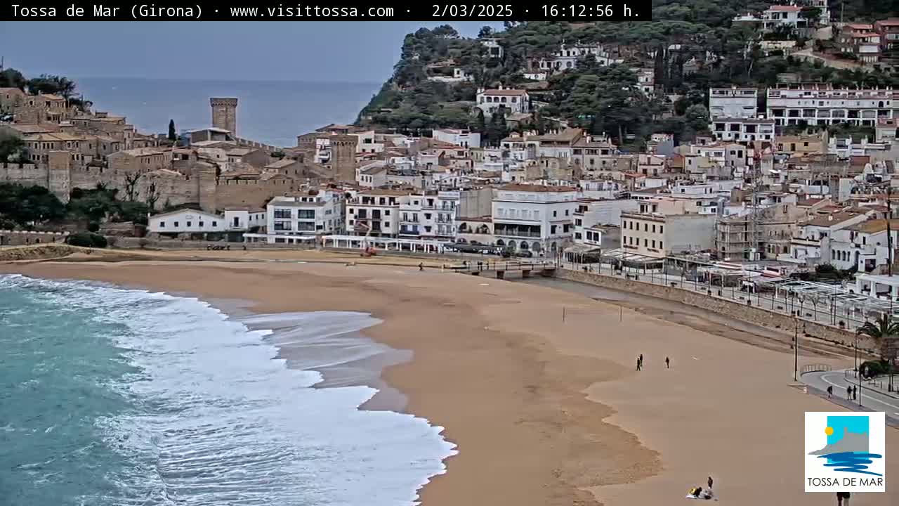 Waves crash on a sandy beach in front of a coastal town with buildings cascading down a hillside.