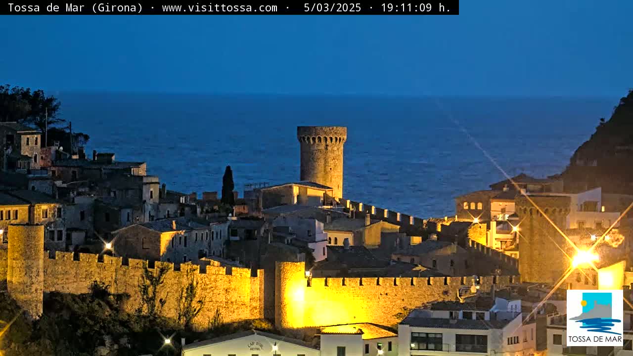 A walled medieval town with a prominent tower is illuminated at night overlooking a dark blue sea.