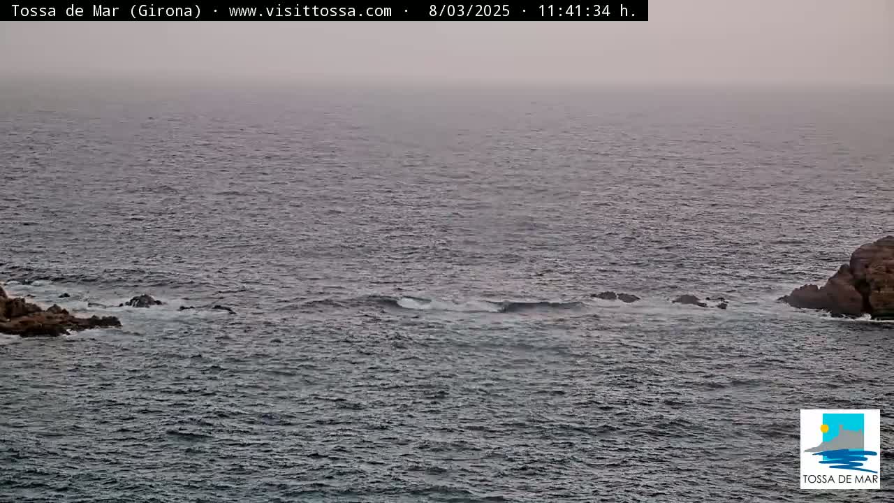 A hazy ocean scene shows dark-colored water with small waves breaking against rocky outcrops on either side of a channel.