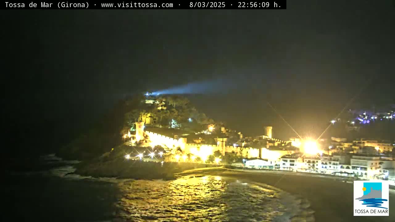 A nighttime coastal scene shows a hilltop castle and town illuminated, with calm ocean waves gently lapping the shore.