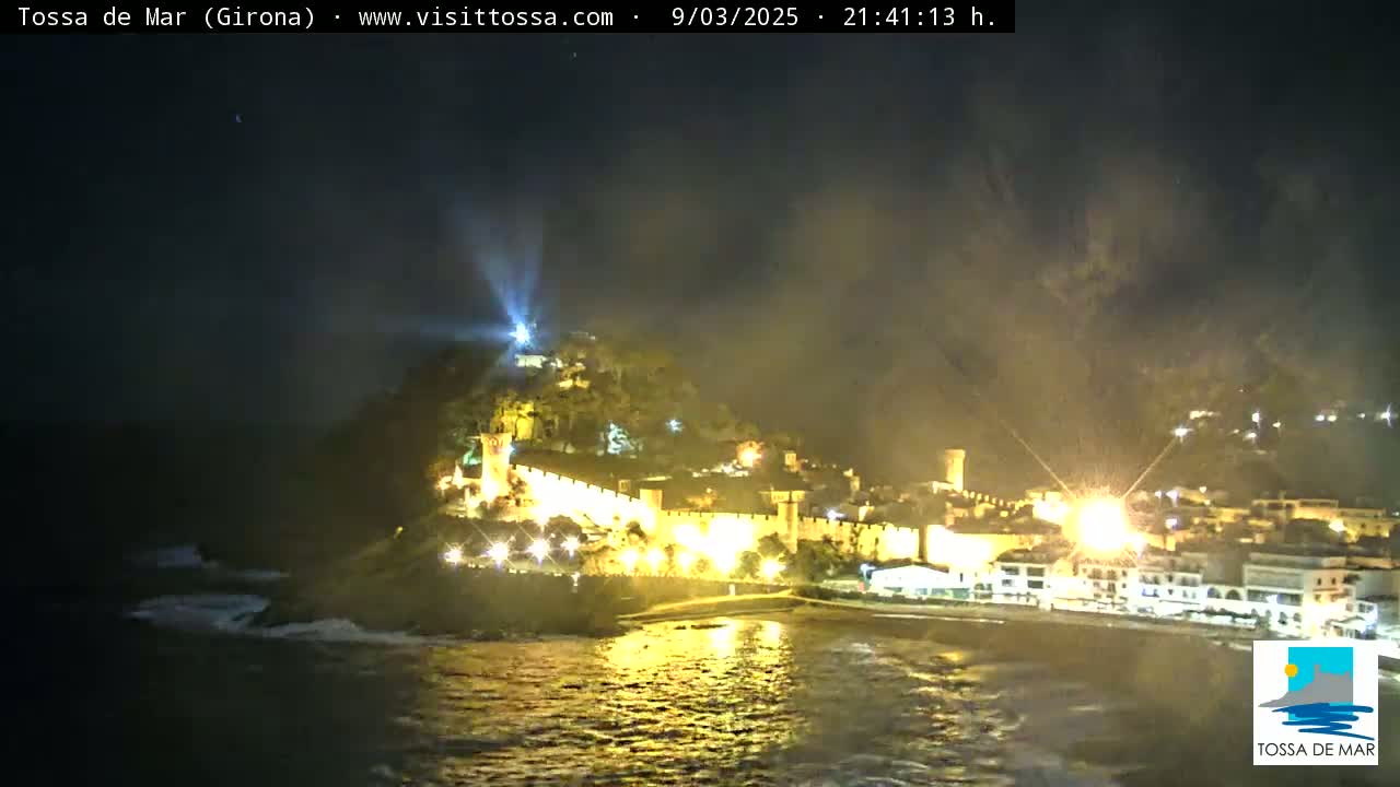 A nighttime coastal scene shows a hilltop town illuminated with lights, partially obscured by a hazy atmosphere, overlooking a dark ocean with small waves.