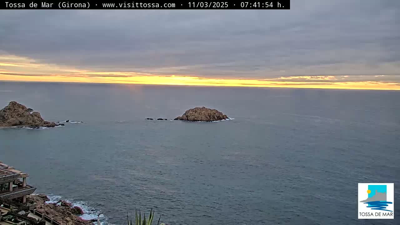 A sunrise over a calm ocean with two rocky islands and part of a building visible on the left.