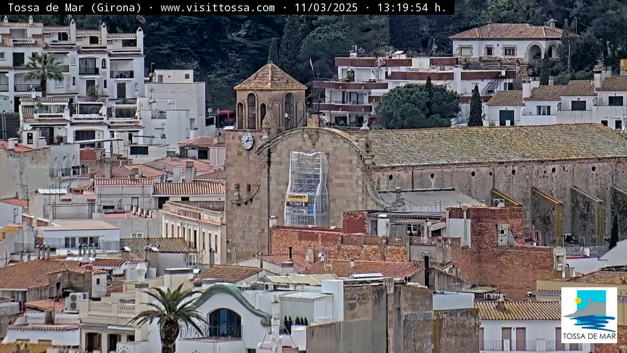 A stone church undergoing renovations is nestled among a dense collection of buildings on a hillside under a mostly clear sky.