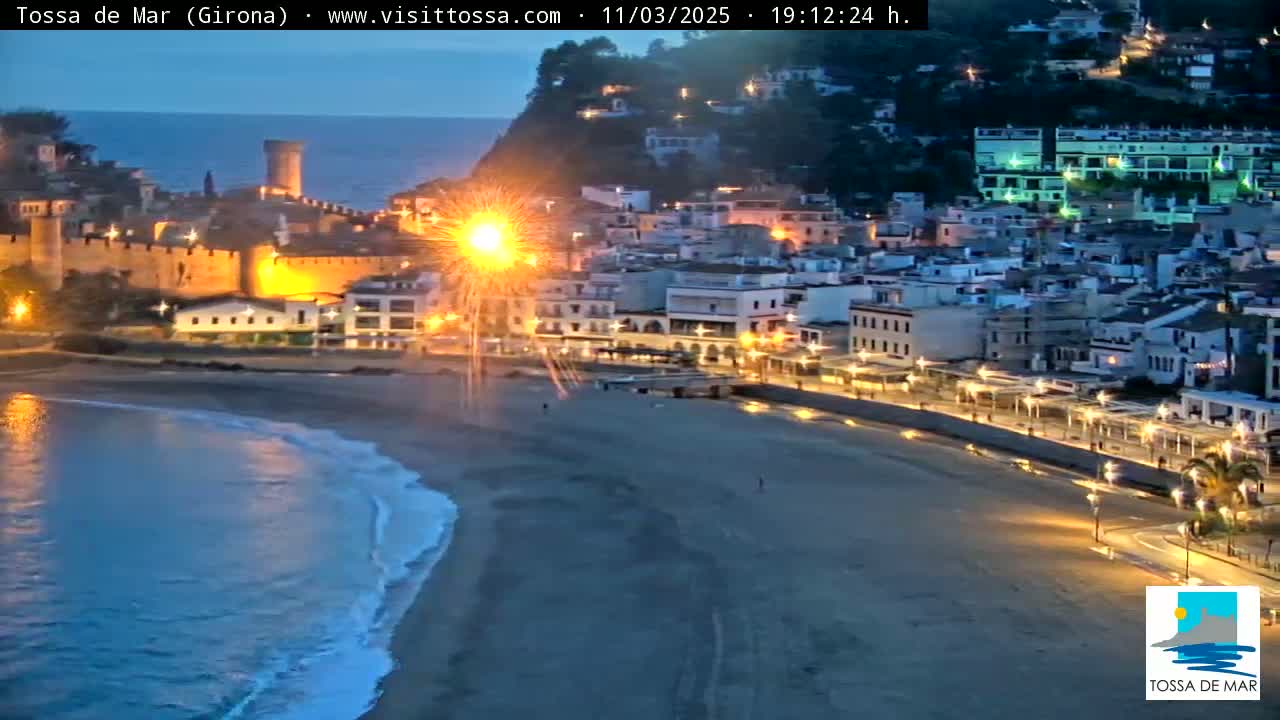 A nighttime coastal town with a medieval castle is illuminated, featuring fireworks over a beach with gentle waves under a twilight sky.