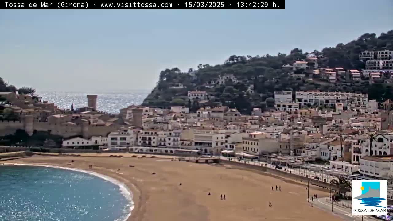 A coastal town with a sandy beach and clear blue water sits beneath a hill covered in vegetation and buildings under a sunny sky.