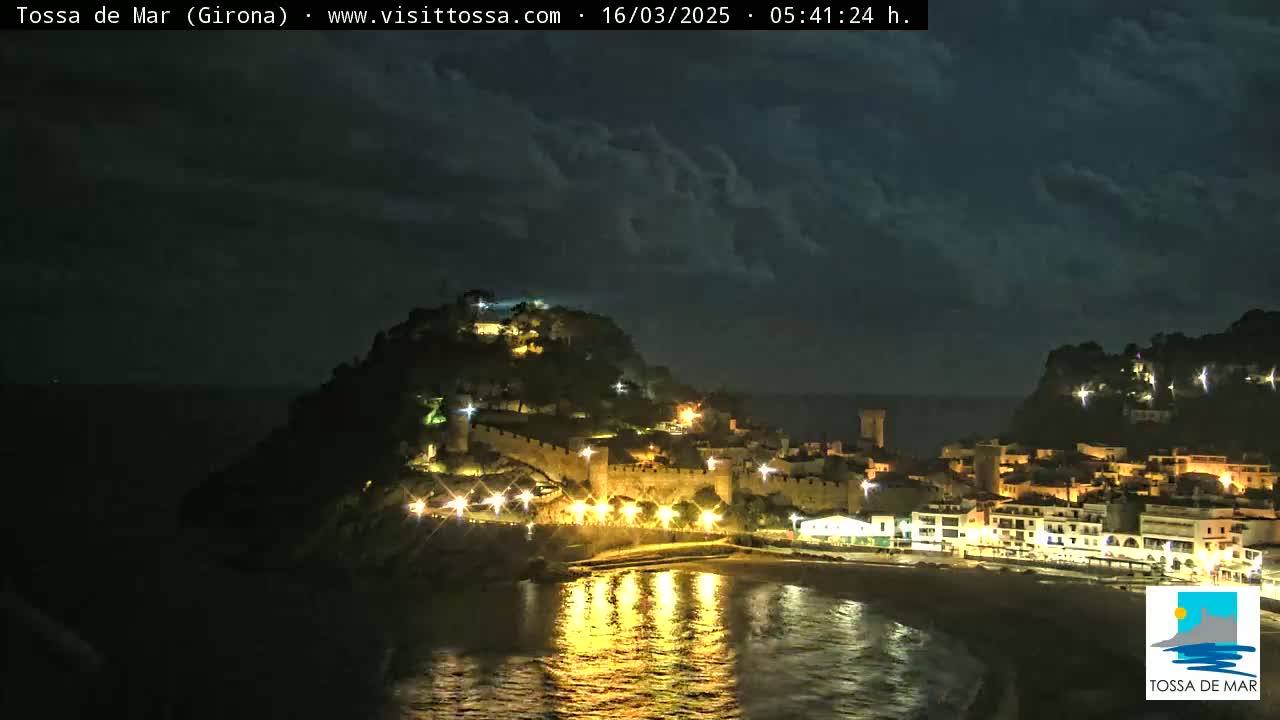 A nighttime coastal town, illuminated with lights, sits beneath a partly cloudy sky.
