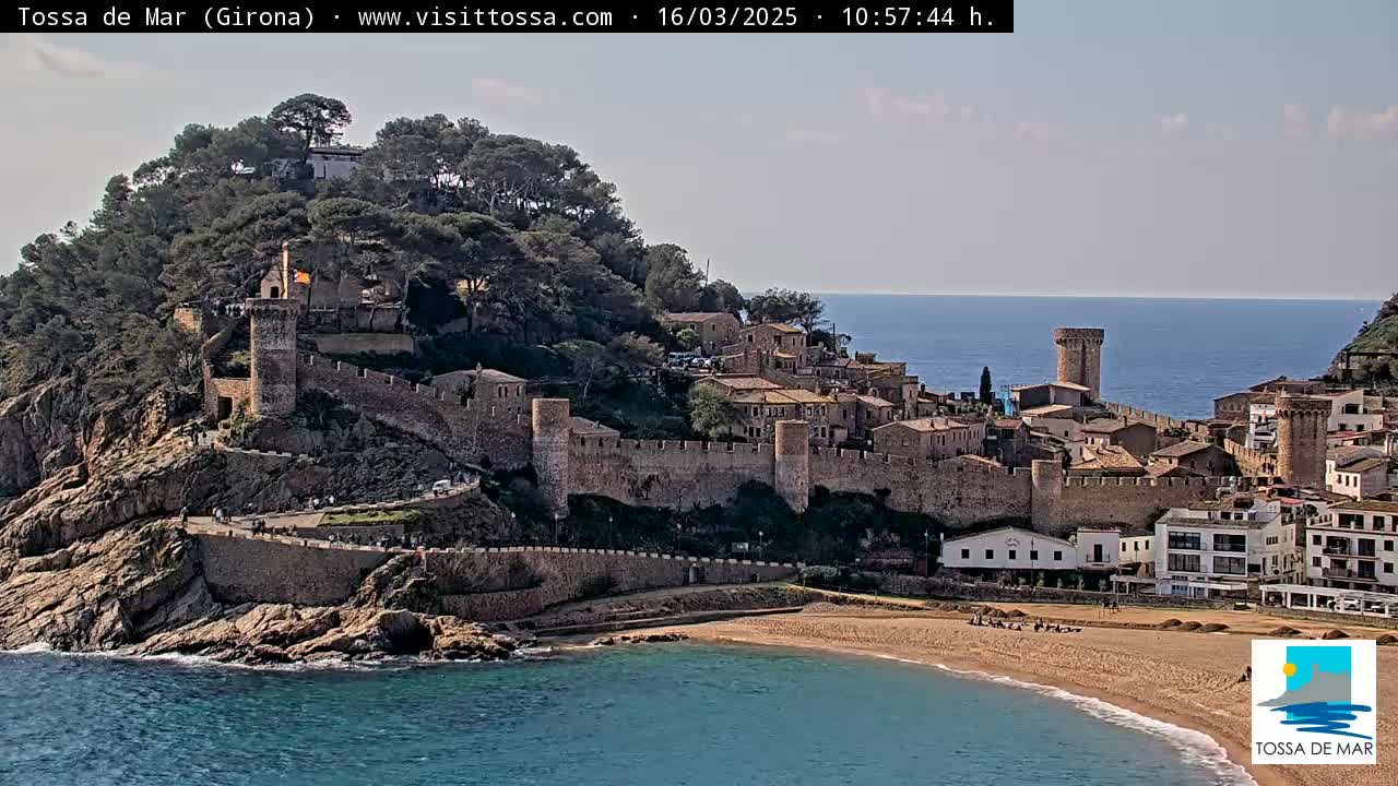 A coastal town with stone walls and towers sits on a rocky hill overlooking a sandy beach and calm blue sea under a partly cloudy sky.