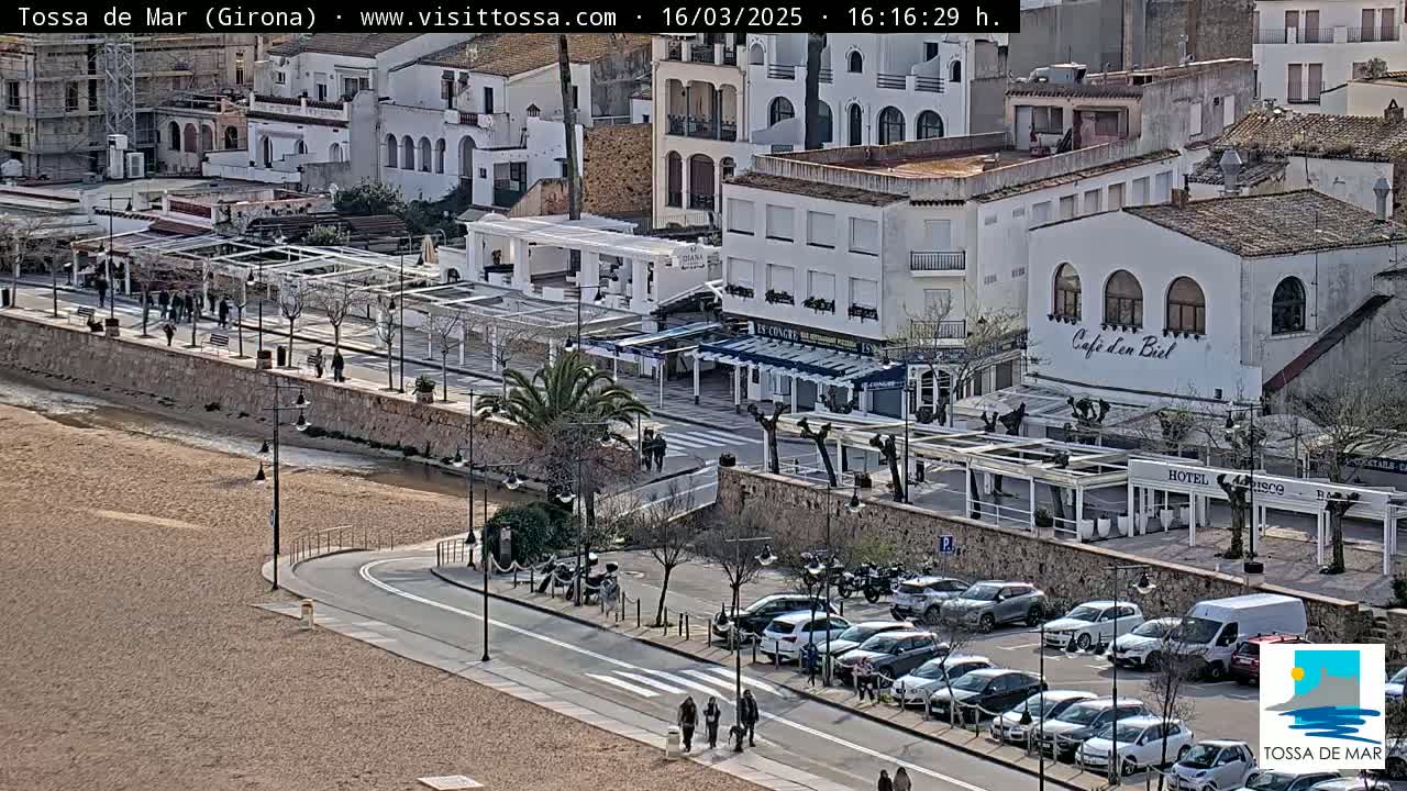 A sunny coastal town scene shows a sandy beach, a paved road curving along the shore, white buildings with outdoor cafes, and a parking lot filled with cars.