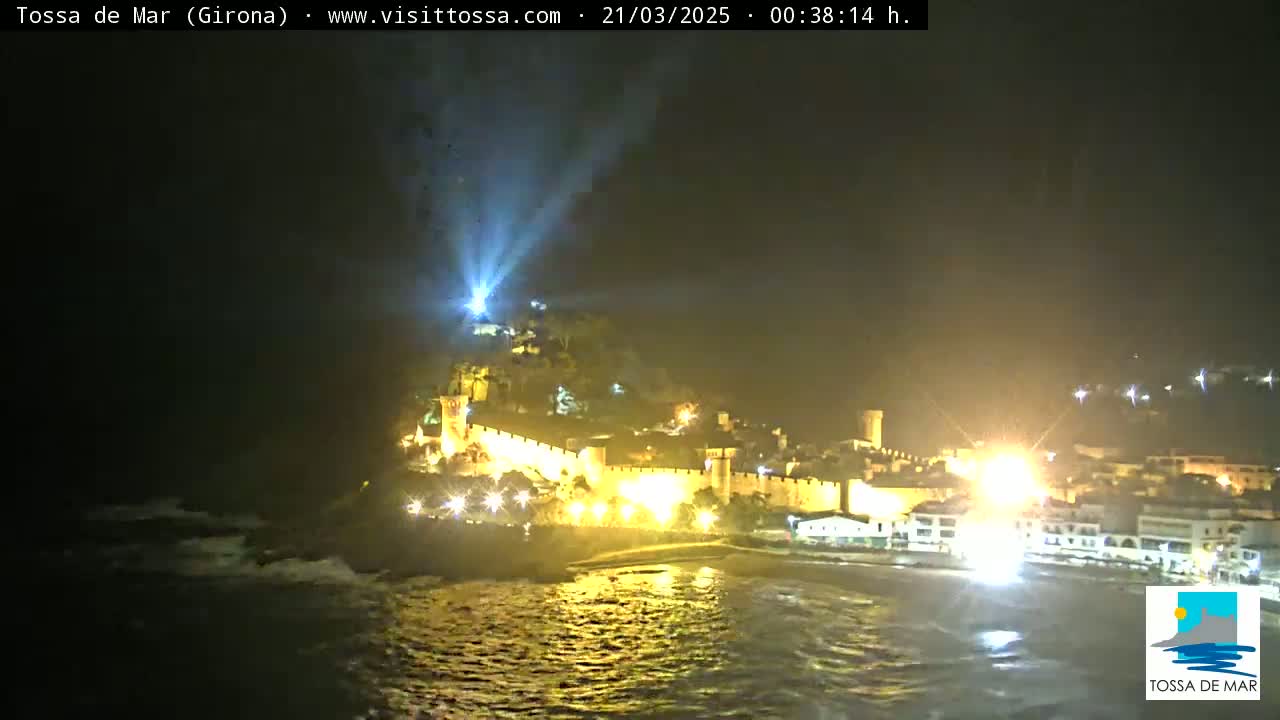 A nighttime coastal scene shows a walled town brightly lit, with a spotlight beam shining upwards from below, and slightly choppy water in the foreground.