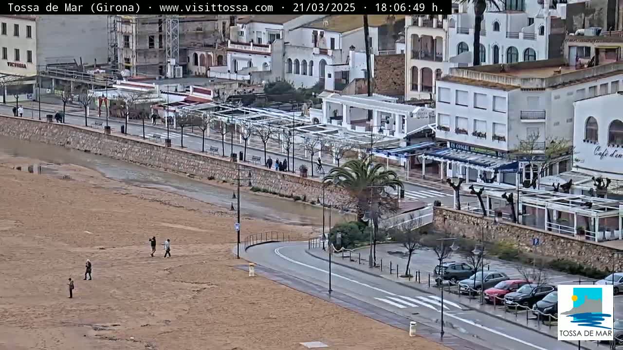 A sandy beach with a few people walking on it is next to a paved road lined with buildings and palm trees, with a waterway running between the beach and the road, on an overcast day.