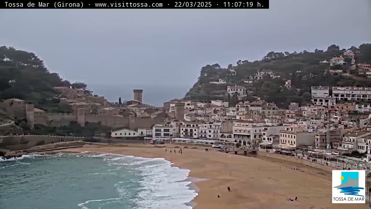 A coastal town with a medieval castle overlooks a sandy beach with moderate waves under an overcast sky.