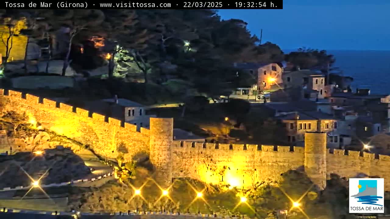A nighttime view of a coastal town's illuminated medieval walls and buildings overlooking a dark sea under a twilight sky.
